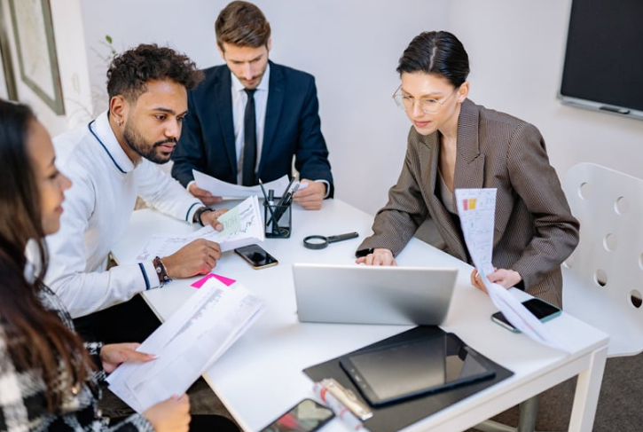 Business team members reviewing documents and discussing project details together during a meeting around a laptop in a modern office.